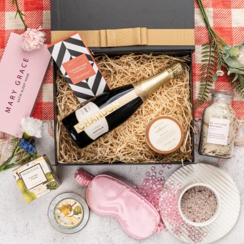 In this carefully composed flat-lay photograph, props of varying sizes and shapes are placed with precision, forming a beautiful frame for a gift hamper. Unbox the gift hamper to reveal a world of delightful items, each one chosen to create a memorable experience. This gift hamper contains: Sleeping Mask; Skincare; Candles; Sparkling Wine; Chocolate; Bath And Shower Gel