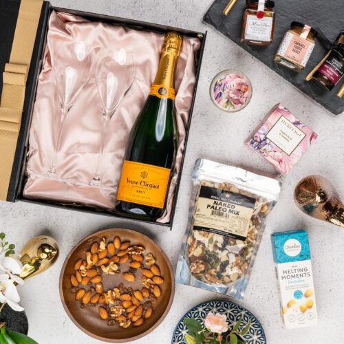 This captivating flat-lay composition entices the viewer with a carefully arranged assortment of props. Inside the gift hamper, you'll find a curated collection of wonderful items, each one chosen to bring joy and happiness, creating lasting memories and spreading warmth and love, igniting a sense of pure happiness. This gift hamper contains: Antipasto; Sparkling Wine; Nuts; Biscuits; Chocolate; Champagne Flutes ; Desert Sauce; Candles