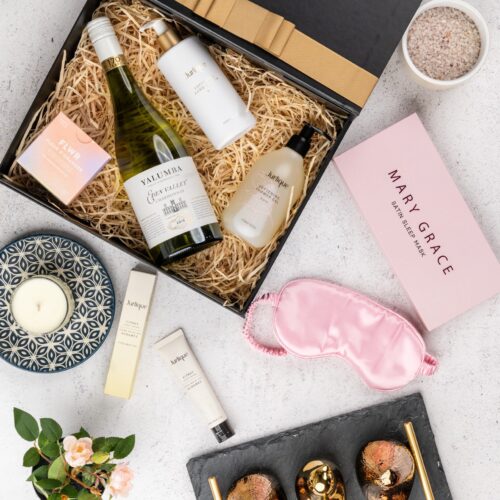 A harmonious blend of textures and colors characterizes this captivating flat-lay photograph, as props converge around a gift hamper. Inside the gift hamper, you'll discover a carefully curated collection of exquisite items, each one a testament to luxury and elegance. This gift hamper contains: White Wine; Sleeping Mask; Candles; Chocolate; Hand Cream