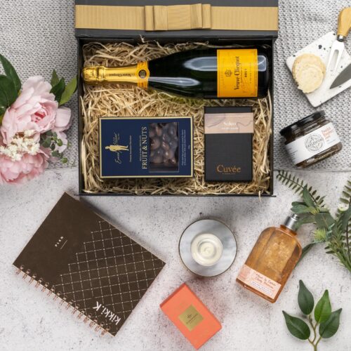 A meticulously arranged collage of props forms a picturesque backdrop for this flat-lay image, enhancing the allure of a gift hamper. Inside the gift hamper, you'll discover a carefully curated selection of treasures. This gift hamper contains: Jam; Chocolate; Sparkling Wine; Candles; Bath And Shower Gel; Planners
