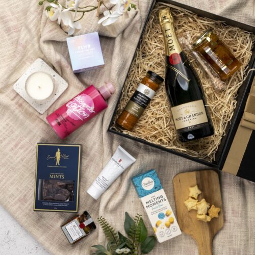 A carefully chosen collection of props forms a visual tapestry around a gift hamper, creating a sense of occasion and celebration. The gift hamper holds an enticing mix of carefully selected products and gourmet delights. This gift hamper contains: Chocolate; Hand Cream; Sparkling Wine; Mustards and Dressings; Candles; Body Lotion; Honey; Antipasto; Biscuits