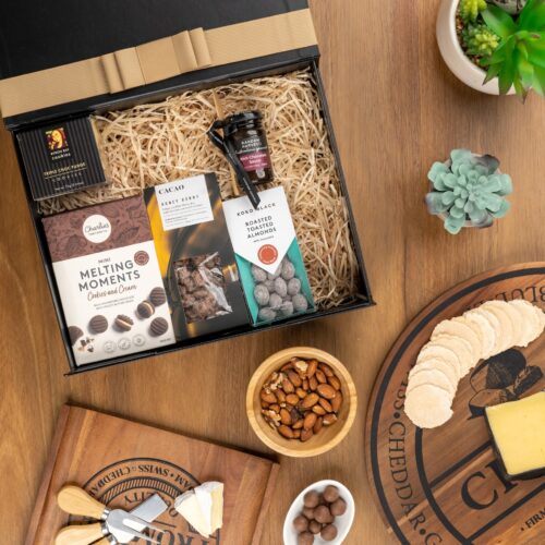 An artful arrangement of props in this captivating flat-lay image creates a sense of anticipation, as they converge towards a central gift hamper. The contents of the gift hamper promise a journey of indulgence and discovery, with each item carefully selected to bring joy. This gift hamper contains: Biscuits; Chocolate