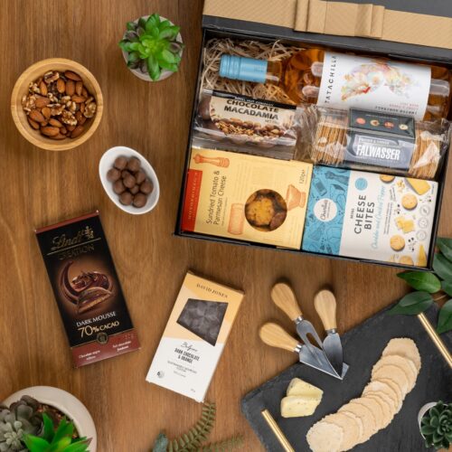 This thoughtfully crafted flat-lay image features an interplay of textures and shapes, as props converge around a gift hamper. Explore the gift hamper and be amazed by the carefully curated assortment of delightful treats and luxurious treasures. This gift hamper contains: Chocolate; Crackers; Biscuits; Rose Wine