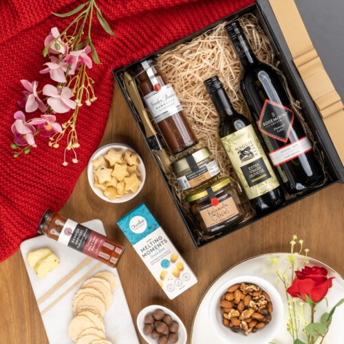 In this visually striking flat-lay image, an assortment of props is skillfully positioned to enhance the allure of a gift hamper. The contents of the gift hamper are a thoughtfully chosen selection of delightful treats and gourmet delicacies. This gift hamper contains: Mustards and Dressings; Oil; Biscuits; Antipasto; Red Wine