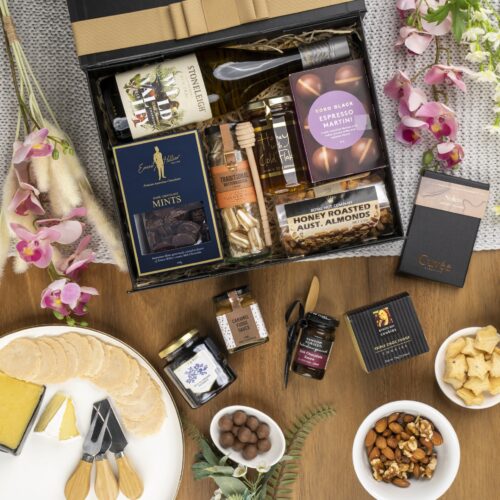In this carefully composed flat-lay photograph, props of varying sizes and shapes are placed with precision, forming a beautiful frame for a gift hamper. Inside the gift hamper, you'll find a curated collection of wonderful items, each one chosen to bring joy and happiness. This gift hamper contains: Desert Sauce; Chocolate; White Wine; Nuts; Jam; Honey; Biscuits