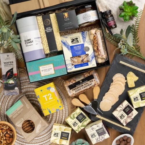 An artful arrangement of props in this captivating flat-lay image creates a sense of anticipation, as they converge towards a central gift hamper. The contents of the gift hamper are a carefully chosen selection of delightful treats and luxurious surprises. This gift hamper contains: Biscuits; Jam; Tea (Black); Nuts; Chocolate; Chutney and Pastes; Crackers