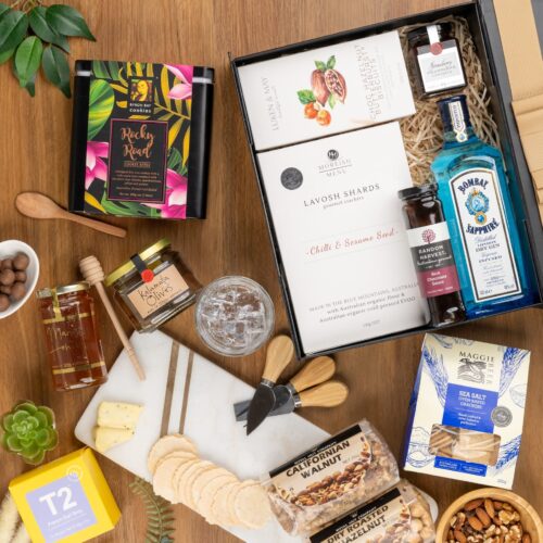 An artful arrangement of props in this captivating flat-lay image creates a sense of anticipation, as they converge towards a central gift hamper. Unveil the gift hamper to reveal a carefully selected assortment of delights, designed to captivate and impress. This gift hamper contains: Nuts; Jam; Biscuits; Crackers; Gin; Desert Sauce; Antipasto; Tea (Black); Honey