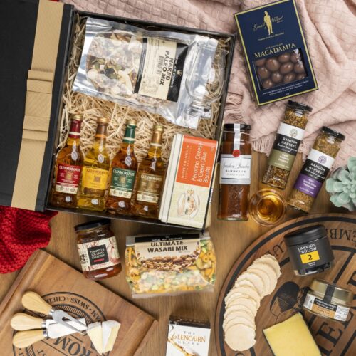 In this visually stunning flat-lay photograph, an assembly of props creates a visual narrative, highlighting the beauty and elegance of a gift hamper. Open the gift hamper to uncover a meticulously chosen selection of items, each one contributing to a truly delightful experience, leaving the recipient in awe and filled with gratitude. This gift hamper contains: Whisky; Chutney and Pastes; Chocolate; Nuts; Mustards and Dressings; Biscuits
