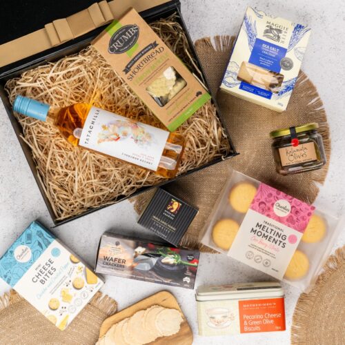 This meticulously styled flat-lay image showcases an array of props artfully positioned around a gift hamper, exuding a sense of excitement and intrigue. Unveil the gift hamper to reveal an assortment of carefully selected delights, guaranteed to impress. This gift hamper contains: Crackers; Biscuits; Antipasto; Rose Wine