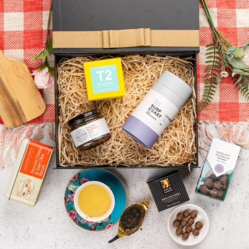 A symphony of colors and textures unfolds in this captivating flat-lay photograph, as an array of props encircle a gift hamper. Inside the gift hamper, you'll find a curated collection of wonderful items, each one chosen to bring joy and happiness, creating lasting memories and spreading warmth and love. This gift hamper contains: Biscuits; Tea (Green); Jam; Chocolate; Teaware; Tea (Black)
