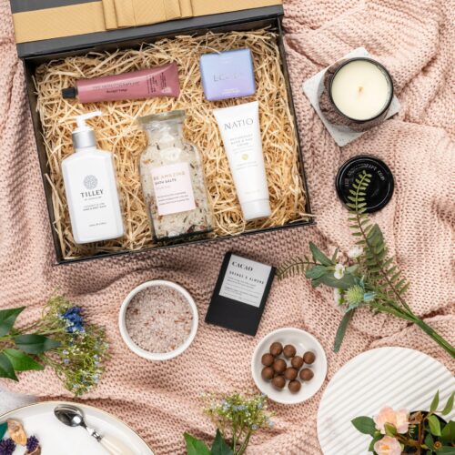 This thoughtfully crafted flat-lay image features an interplay of textures and shapes, as props converge around a gift hamper. Open the gift hamper to uncover a meticulously chosen selection of items, carefully arranged to bring delight. This gift hamper contains: Chocolate; Hand Cream; Candles; Body Wash; Bath And Shower Gel; Soap