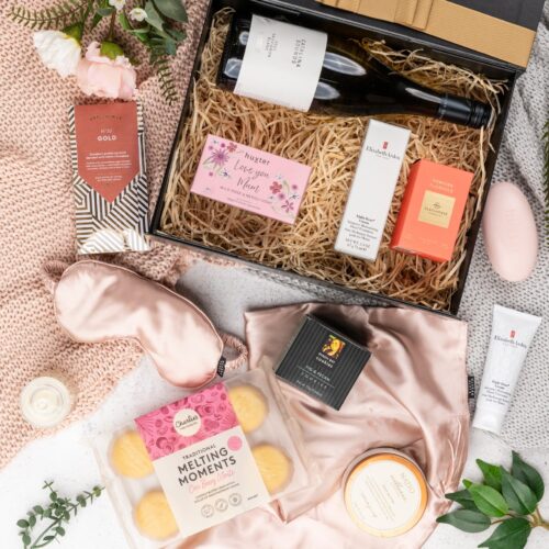 In this mesmerizing flat-lay composition, an assortment of carefully chosen props forms a visual tapestry around a gift hamper. Open the gift hamper to reveal a delightful surprise waiting to be savored, a true feast for the senses. This gift hamper contains: Sleeping Mask; Candles; Biscuits; Skincare; Hand Cream; Chocolate; Soap; White Wine