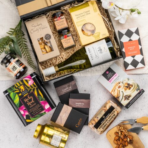 In this visually stunning flat-lay photograph, an assembly of props creates a visual narrative, highlighting the beauty and elegance of a gift hamper. The gift hamper holds an enchanting array of carefully selected surprises, guaranteed to leave a lasting impression. This gift hamper contains: Jam; Antipasto; Crackers; Tea (Black); Chocolate; White Wine; Nuts; Biscuits