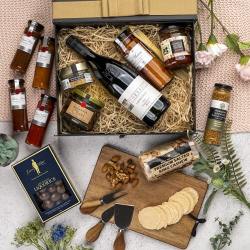 This visually arresting flat-lay image showcases a meticulously styled collection of props, expertly arranged around a gift hamper. Open the gift hamper to uncover a collection of carefully curated treasures and unique finds. This gift hamper contains: Red Wine; Cheese Board; Nuts; Chocolate; Mustards and Dressings; Chutney and Pastes; Antipasto