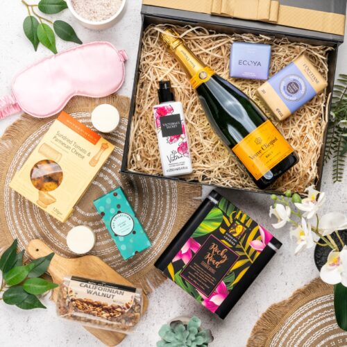 This striking flat-lay image features a curated selection of props that converge around a gift hamper, speaking of generosity and thoughtfulness. Open the gift hamper to reveal a delightful surprise waiting to be savored, each item carefully selected to create a moment of pure bliss and bring a smile to the recipient's face. This gift hamper contains: Sparkling Wine; Nuts; Crackers; Soap; Biscuits; Sleeping Mask; Body Lotion; Skincare; Candles